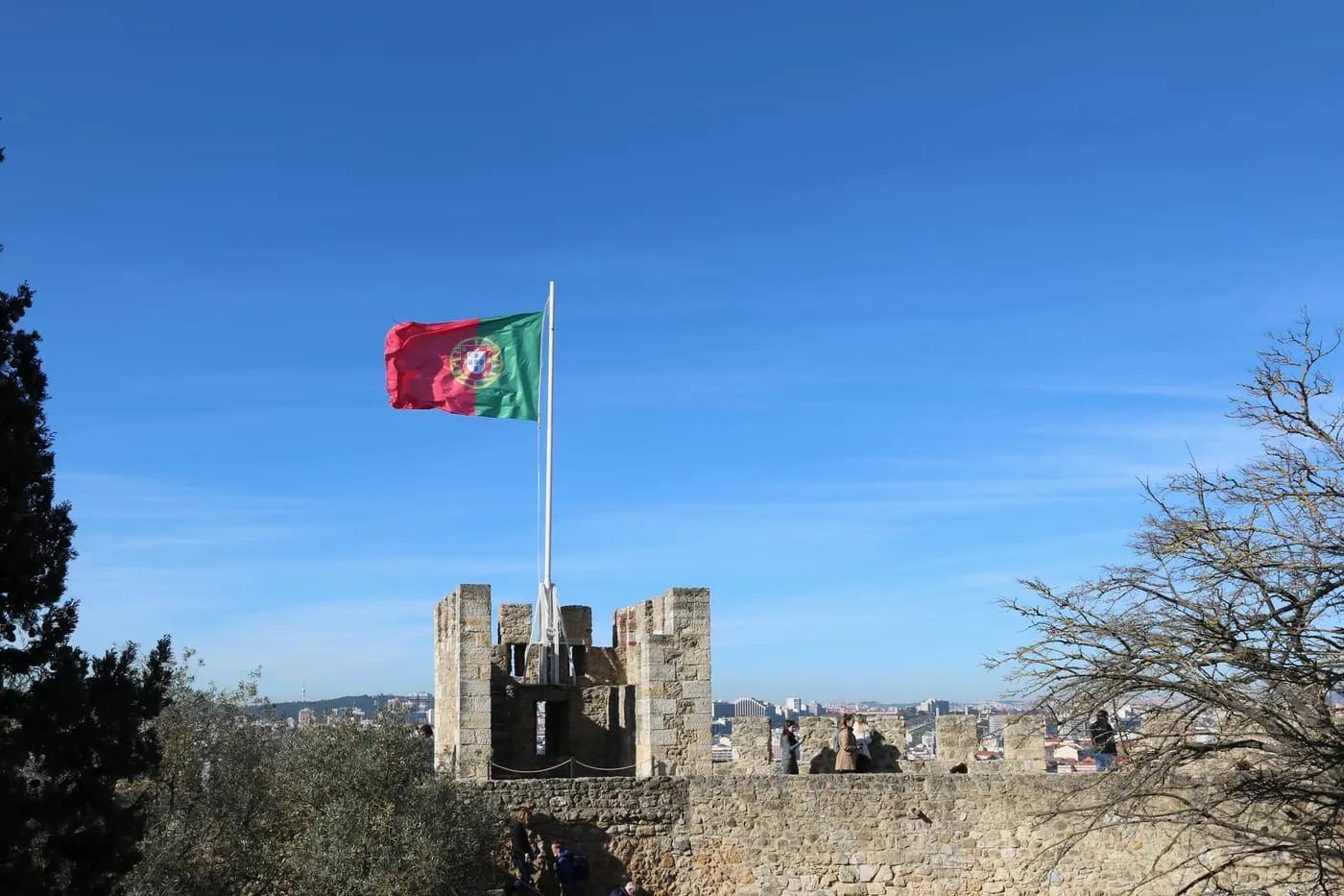 A crenellated tower of the castle with the Portuguese flag on a pole above it and the city below, under a blue sky