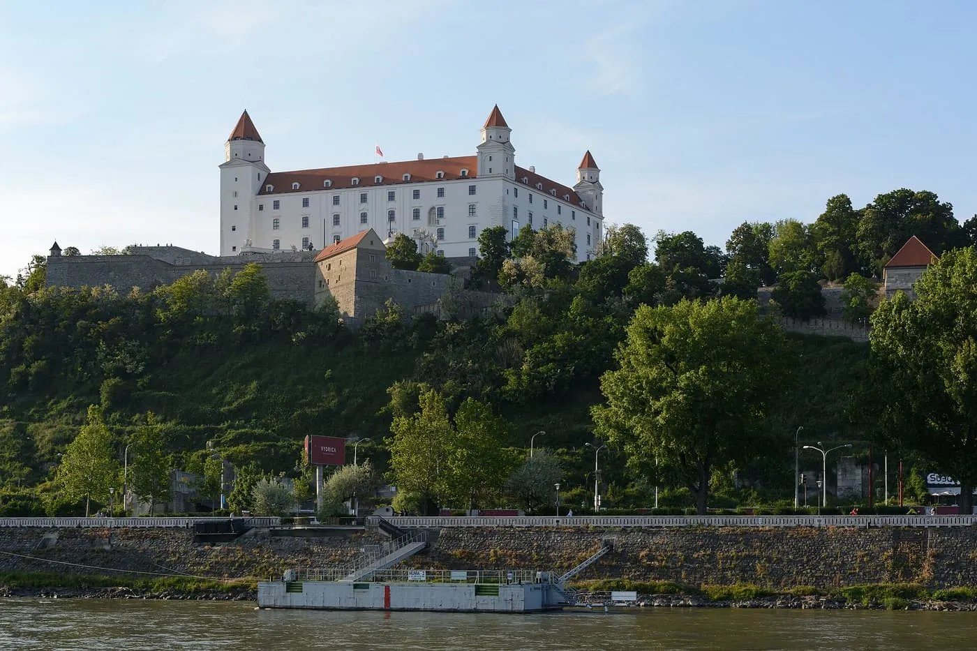 Bratislava Castle, a white block with four corner towers, on a wooded hill above the Danube embankment