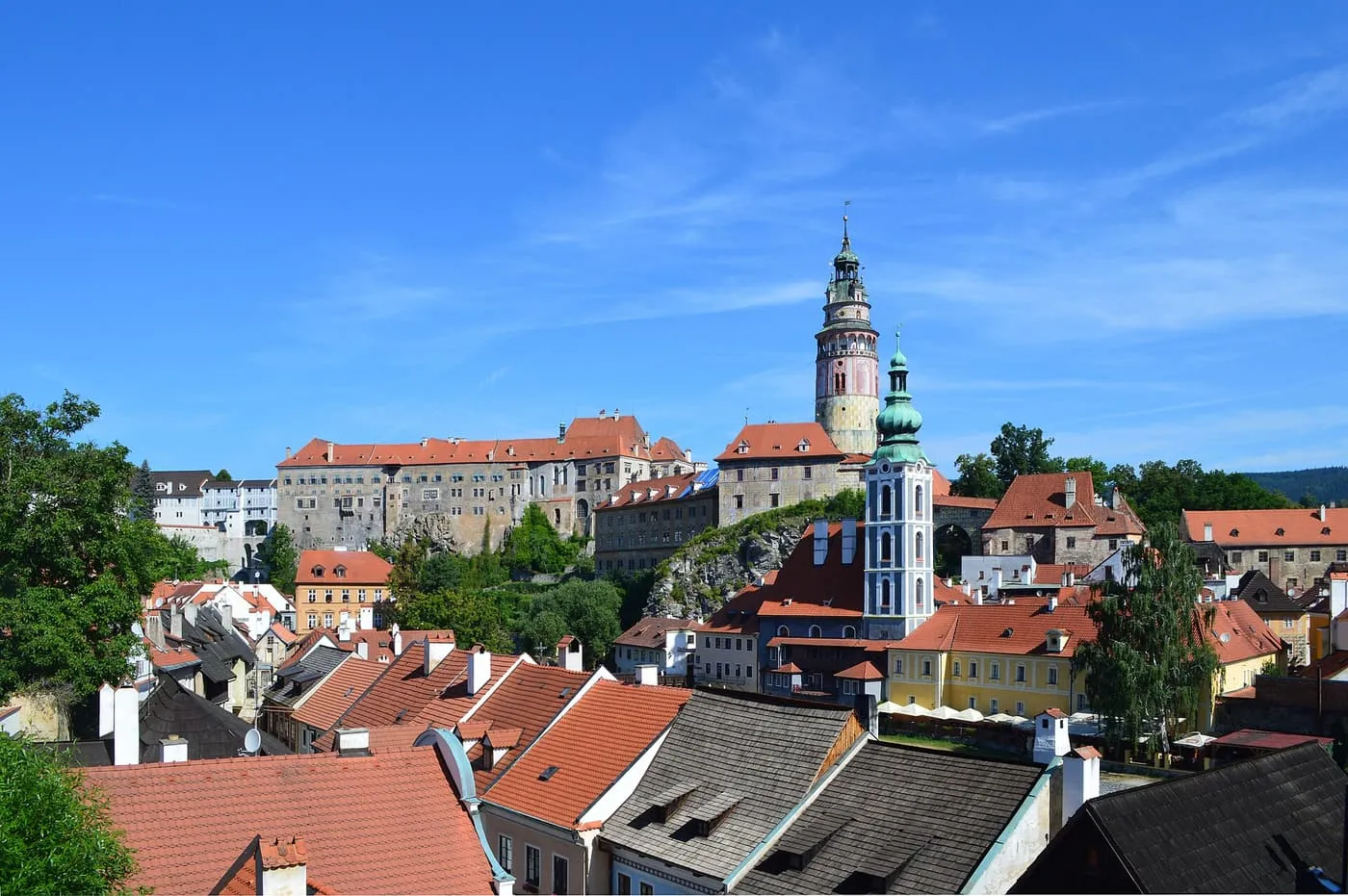 Český Krumlov Castle with its round tower and the green-domed St Jost's church above the red roofs of the old town, under a blue sky