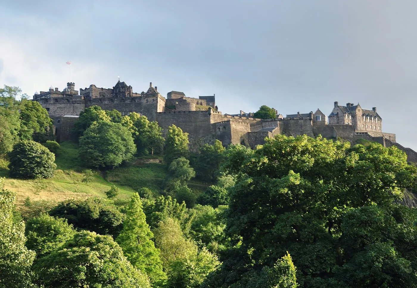 Edinburgh Castle on its crag above the green tree canopy of Princes Street Gardens, under a grey sky with low sun on the walls