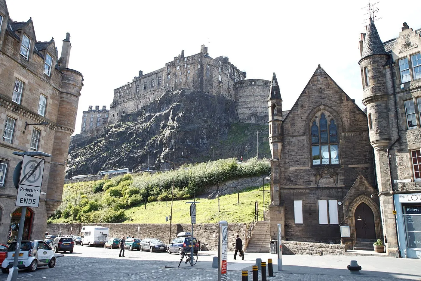 Edinburgh Castle on its crag above a grassy slope and a stone church at the edge of the Grassmarket, with cars parked below