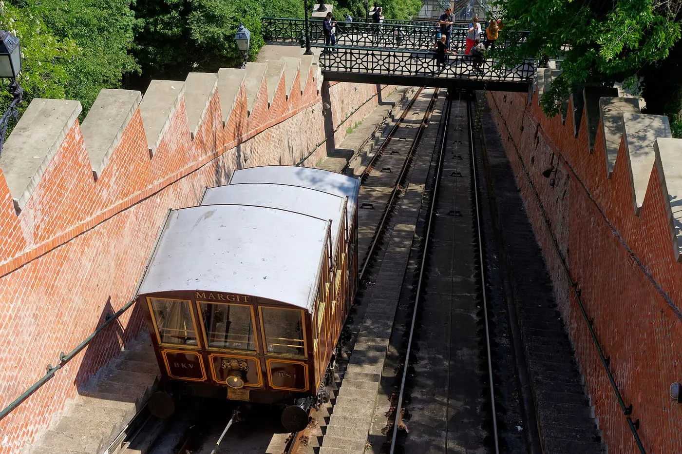 A wooden funicular car on the track between brick walls, with a footbridge above
