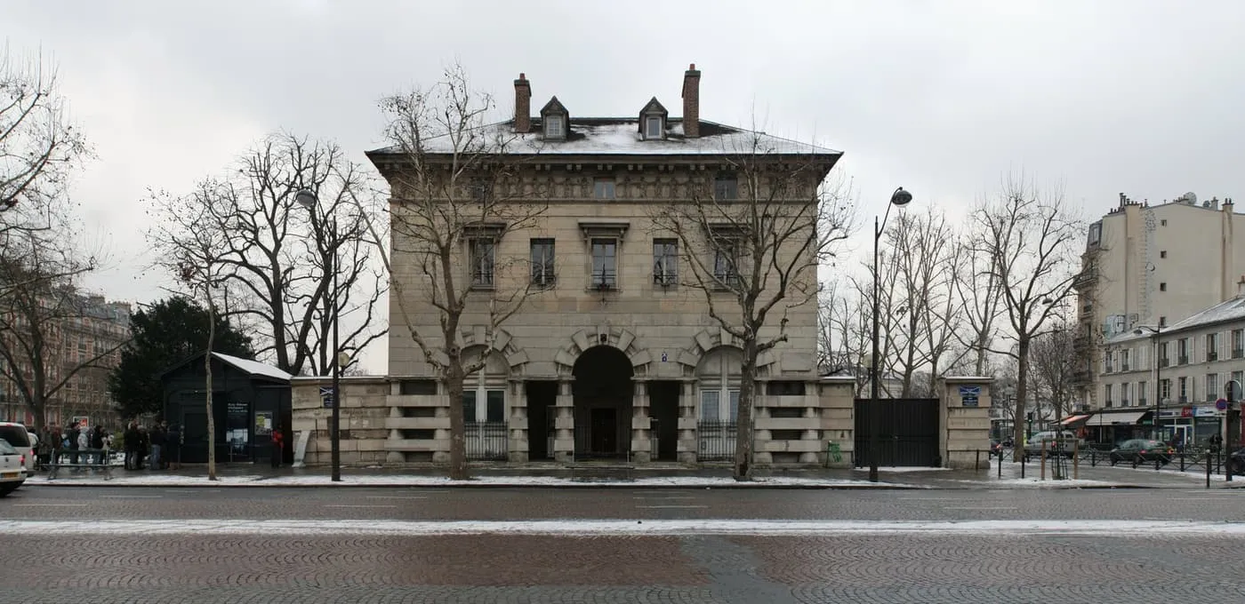 The stone toll house of the Barrière d'Enfer on the Place Denfert-Rochereau, with the dark green Catacombs entrance kiosk at the left edge and a small queue beside it