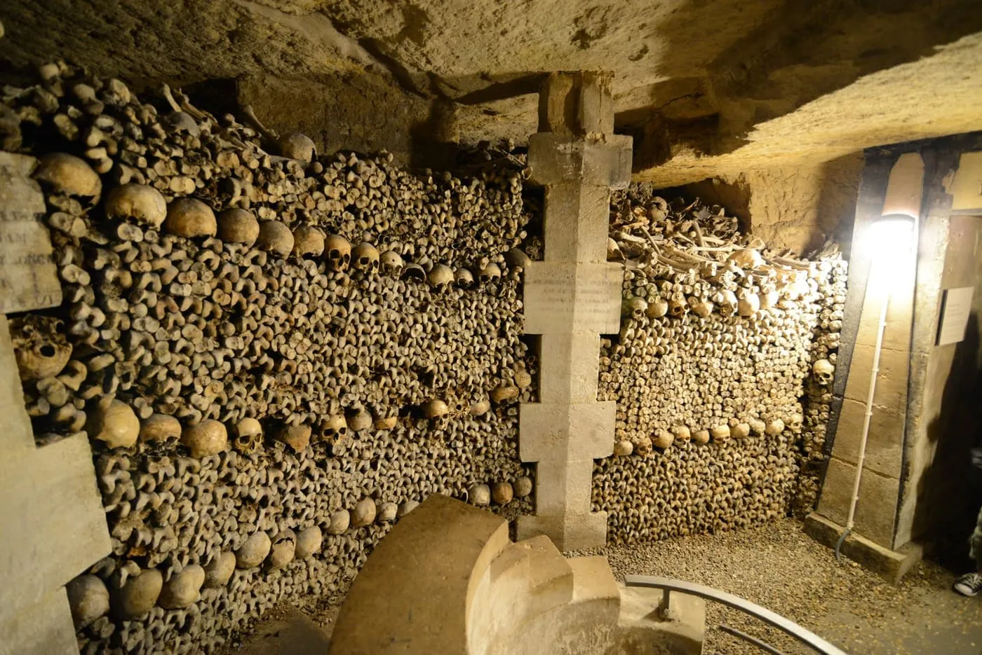 Walls of stacked skulls and thigh bones in the Paris Catacombs ossuary, with a stone pillar between two bays and a lamp on the right