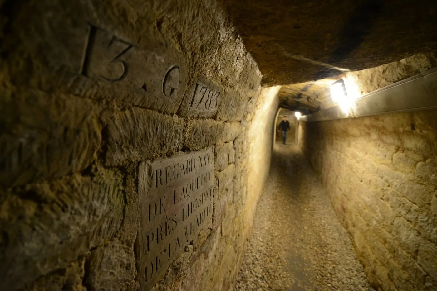 A narrow lamp-lit quarry gallery in the Catacombs, with a dated inscription and a carved plaque on the stone wall and a visitor far down the passage