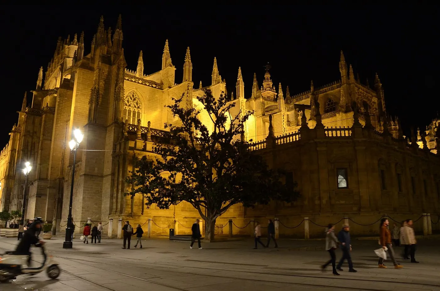 The floodlit Gothic Cathedral of Seville at night, its pinnacles and buttresses lit gold, with pedestrians and a scooter on the paving in front