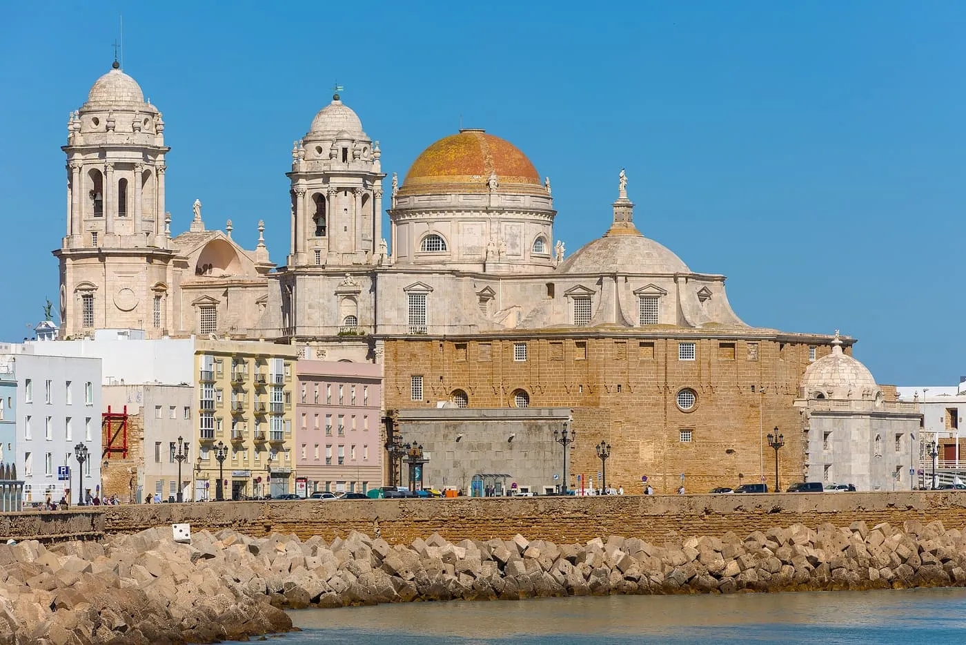 Cádiz Cathedral from the seafront, with its two bell towers, the yellow-tiled dome and the stone flank of the nave, and breakwater rocks in front
