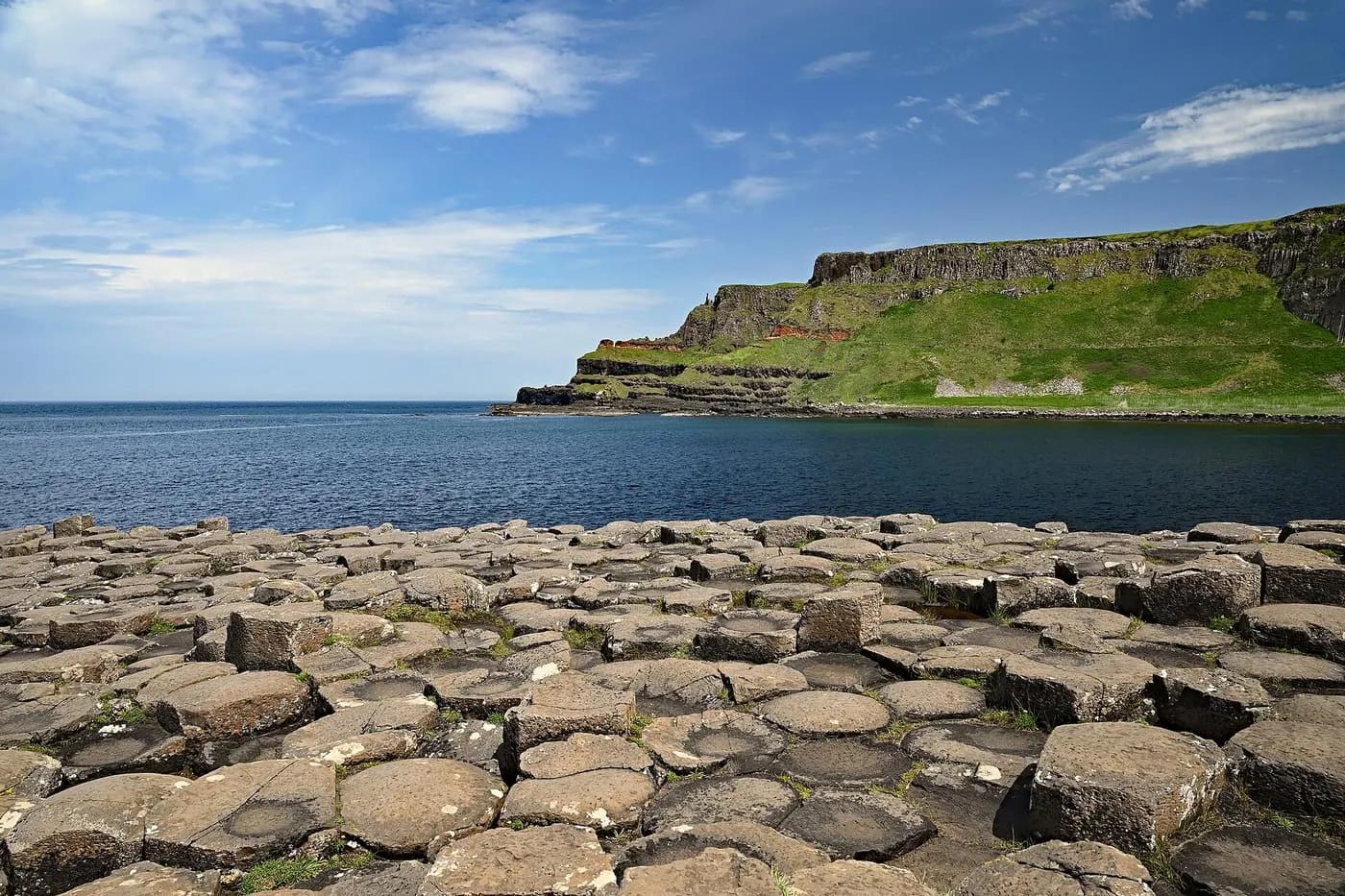Hexagonal basalt stone tops in the foreground, a blue sea inlet and a green headland with dark rock layers beyond