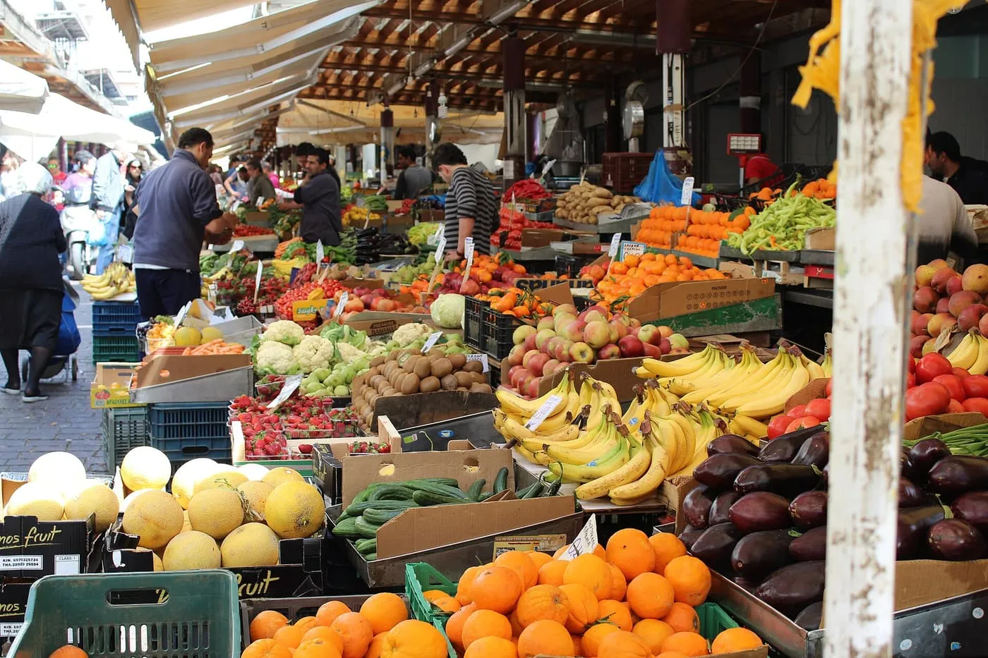 Fruit and vegetable stalls piled with oranges, bananas, aubergines, cauliflowers and strawberries under awnings, with shoppers in the aisle
