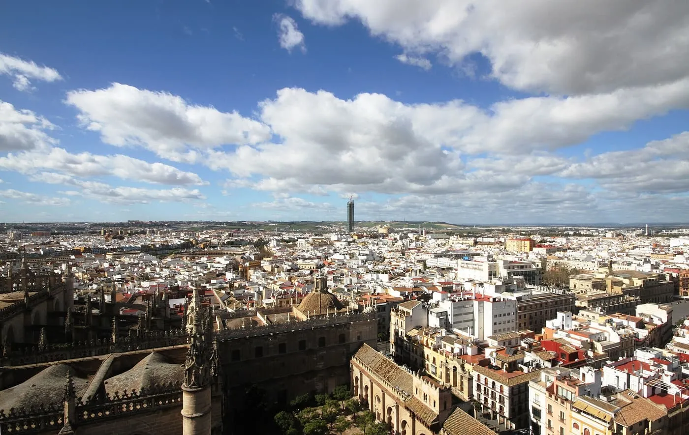 The roofs of the Cathedral and the white rooftops of central Seville seen from above, with a tall tower on the horizon under a cloudy blue sky