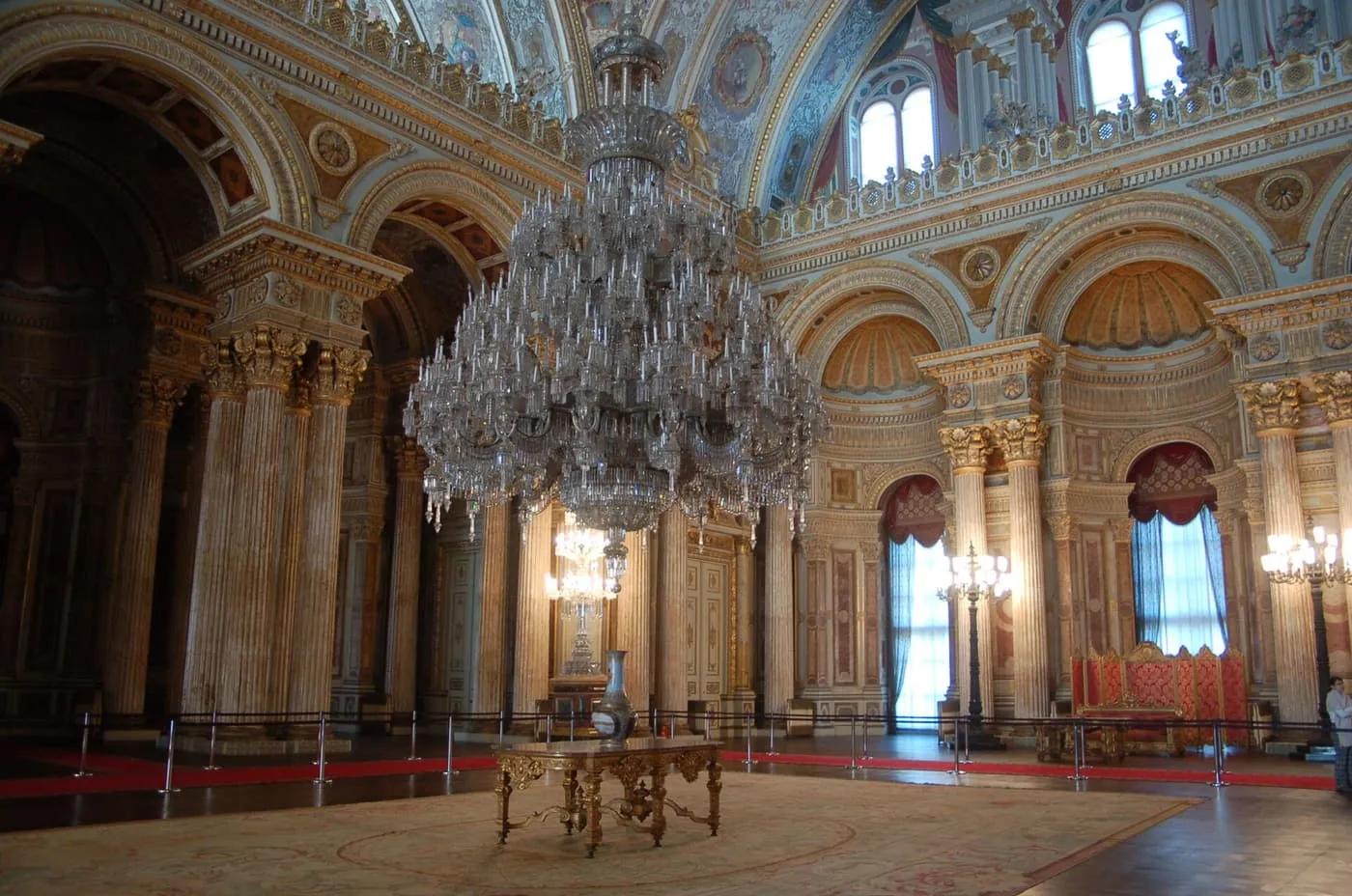 A huge crystal chandelier hanging in a hall of marble columns and arches, with red chairs at the far end
