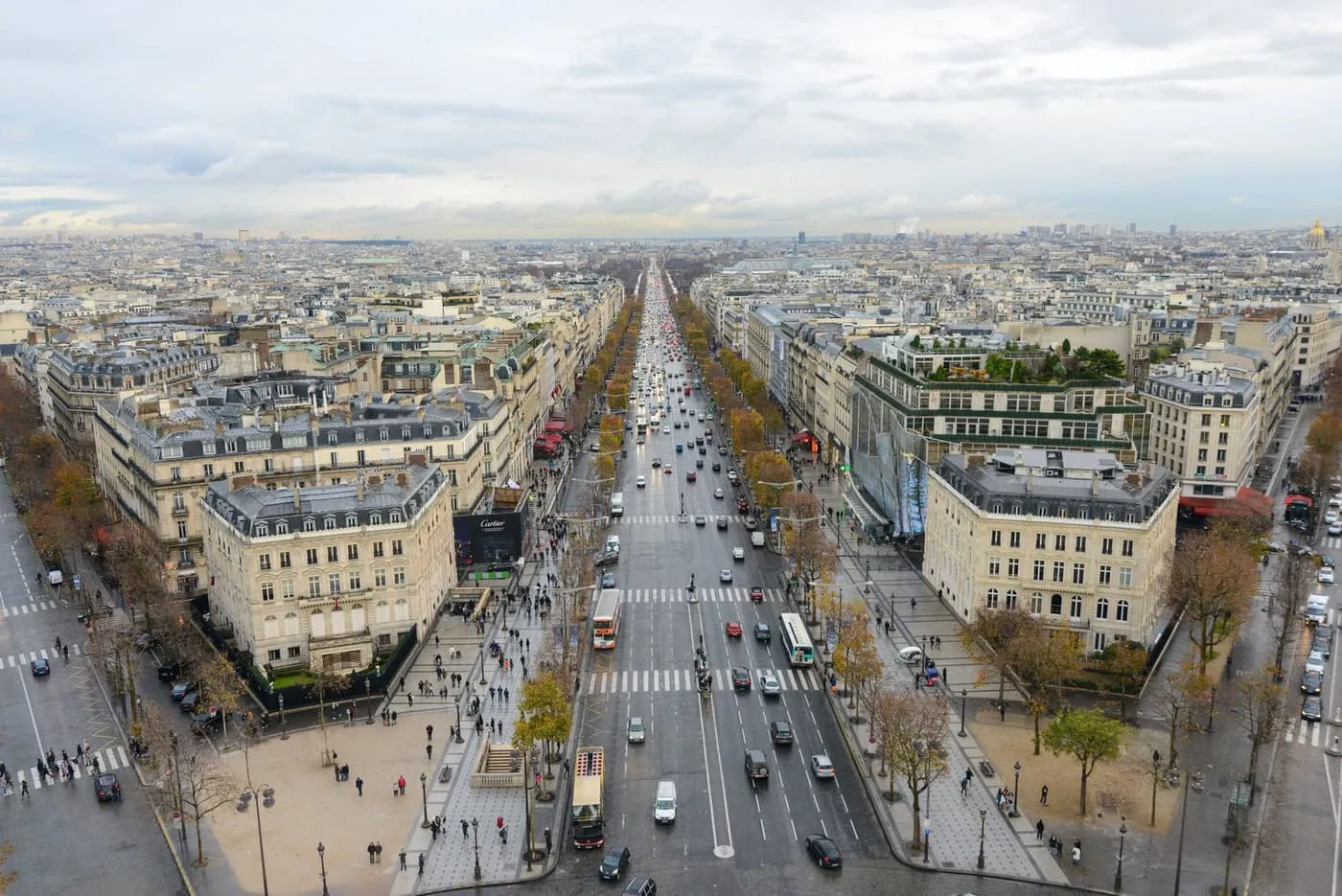 The Champs-Élysées running away from the viewer between Haussmann-era buildings, seen from the roof of the Arc de Triomphe under a cloudy sky