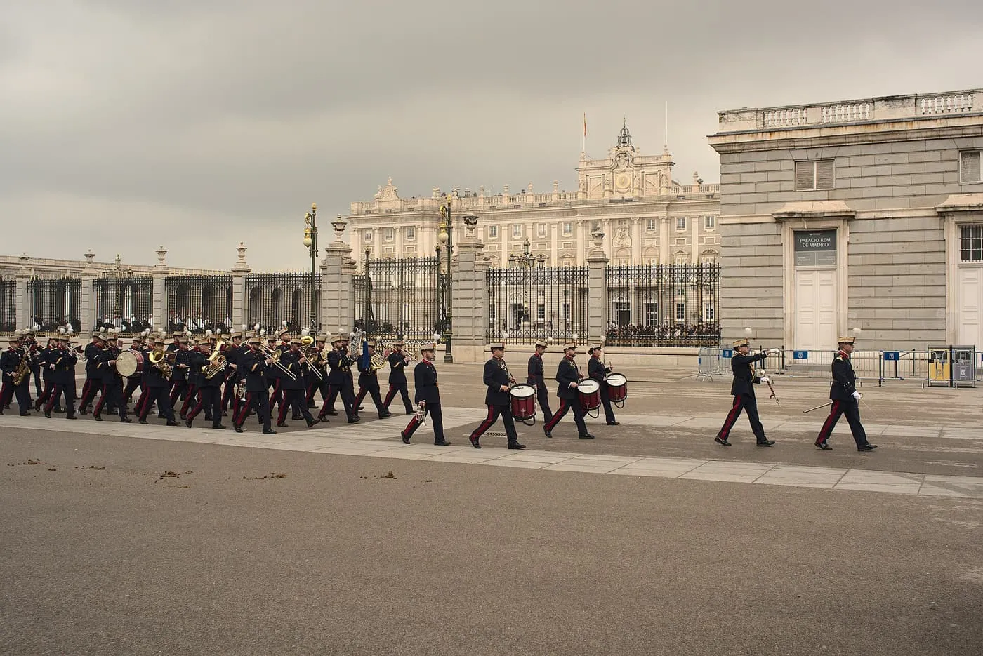 A band of the Royal Guard in dark blue uniforms with red stripes marching across the esplanade in front of the Royal Palace gates