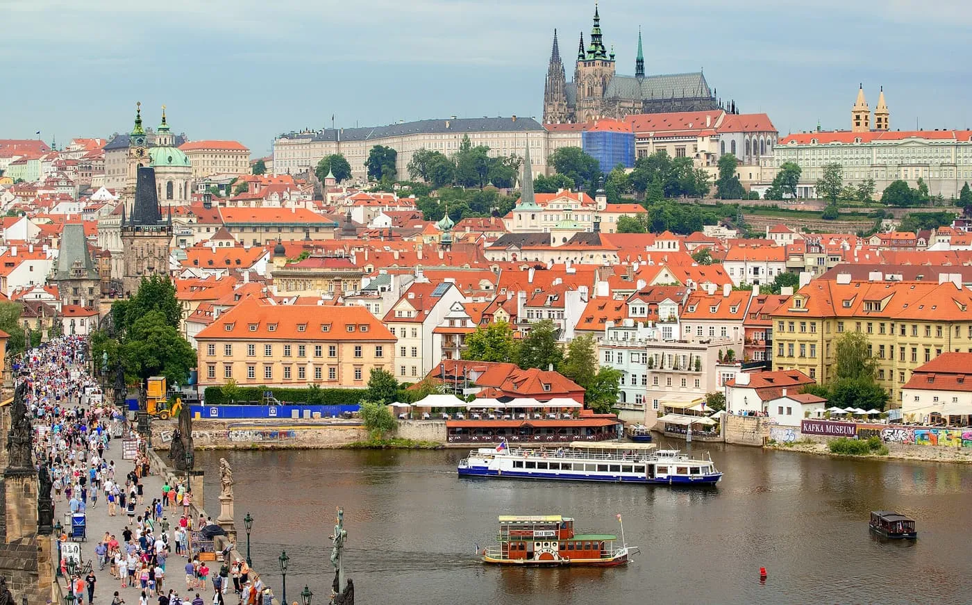 Charles Bridge crowded with walkers, with the red roofs of the Lesser Town below Prague Castle on the hill behind and a river boat on the Vltava