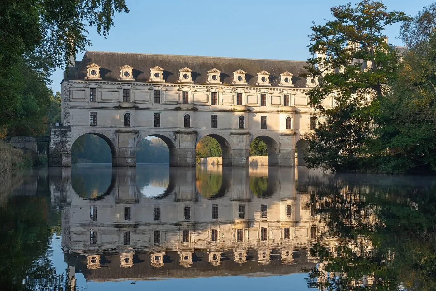 The arched gallery of Château de Chenonceau spanning the River Cher, reflected in still water in morning light