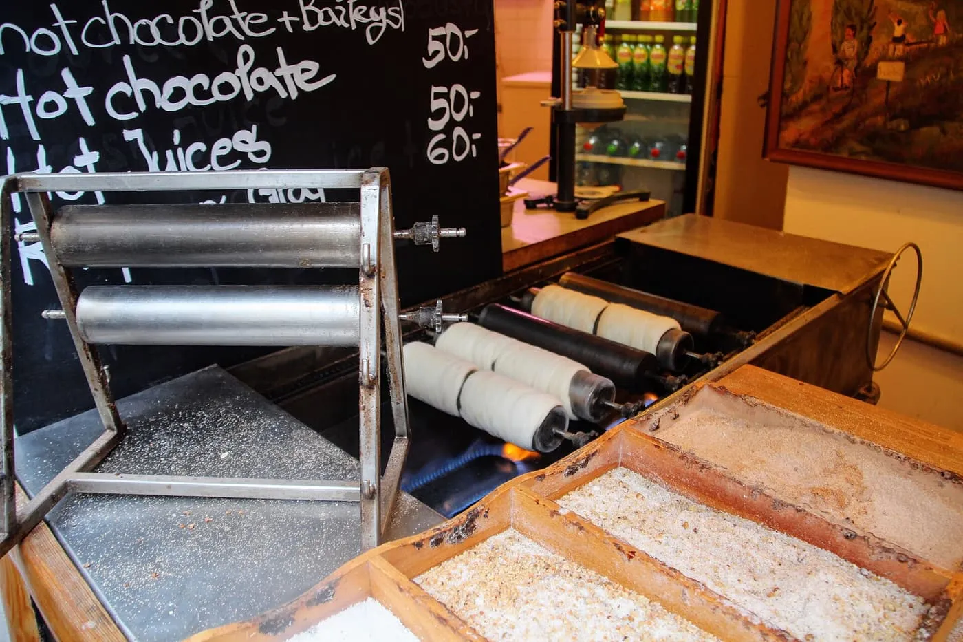 Chimney cakes turning on metal spits over a gas flame at a Prague stall, with trays of sugar and nut coatings in the foreground