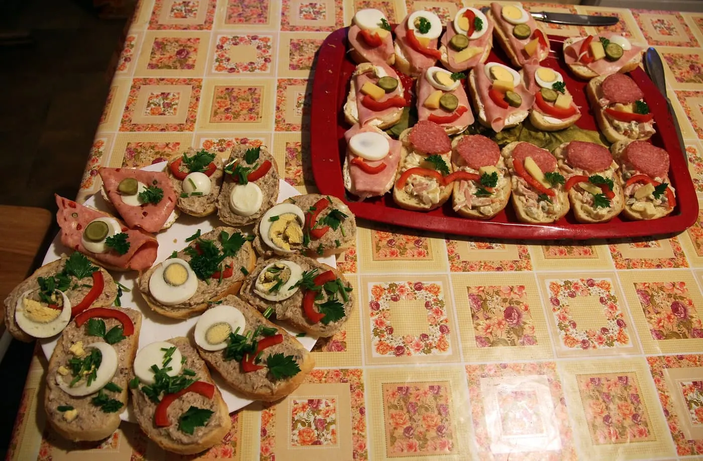 A plate and a tray of chlebíčky, small open sandwiches on sliced baguette topped with ham, salami, egg, pickle and red pepper