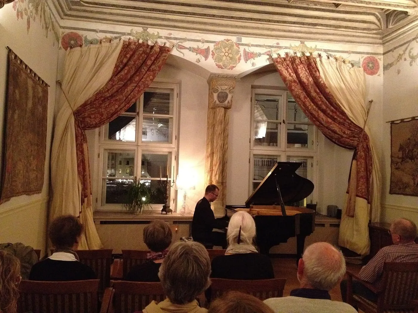A pianist playing a grand piano before an audience in a room with painted ceiling friezes, looped curtains and two tall windows