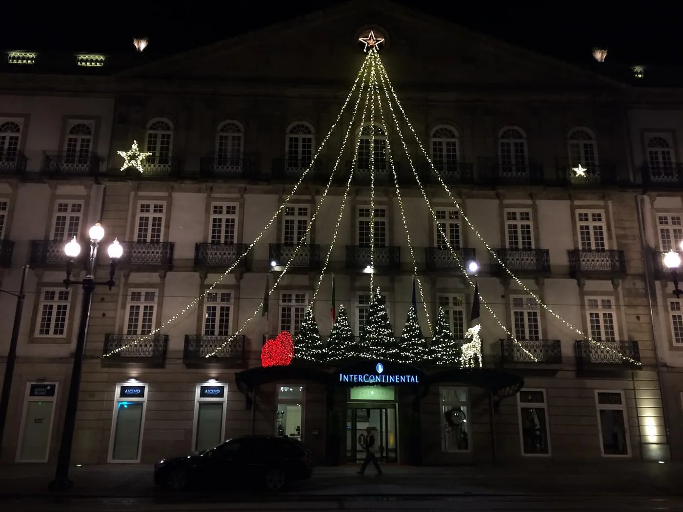 A palace-fronted hotel at night with white lights strung from a star on the roof to the doorway, and lit Christmas trees below