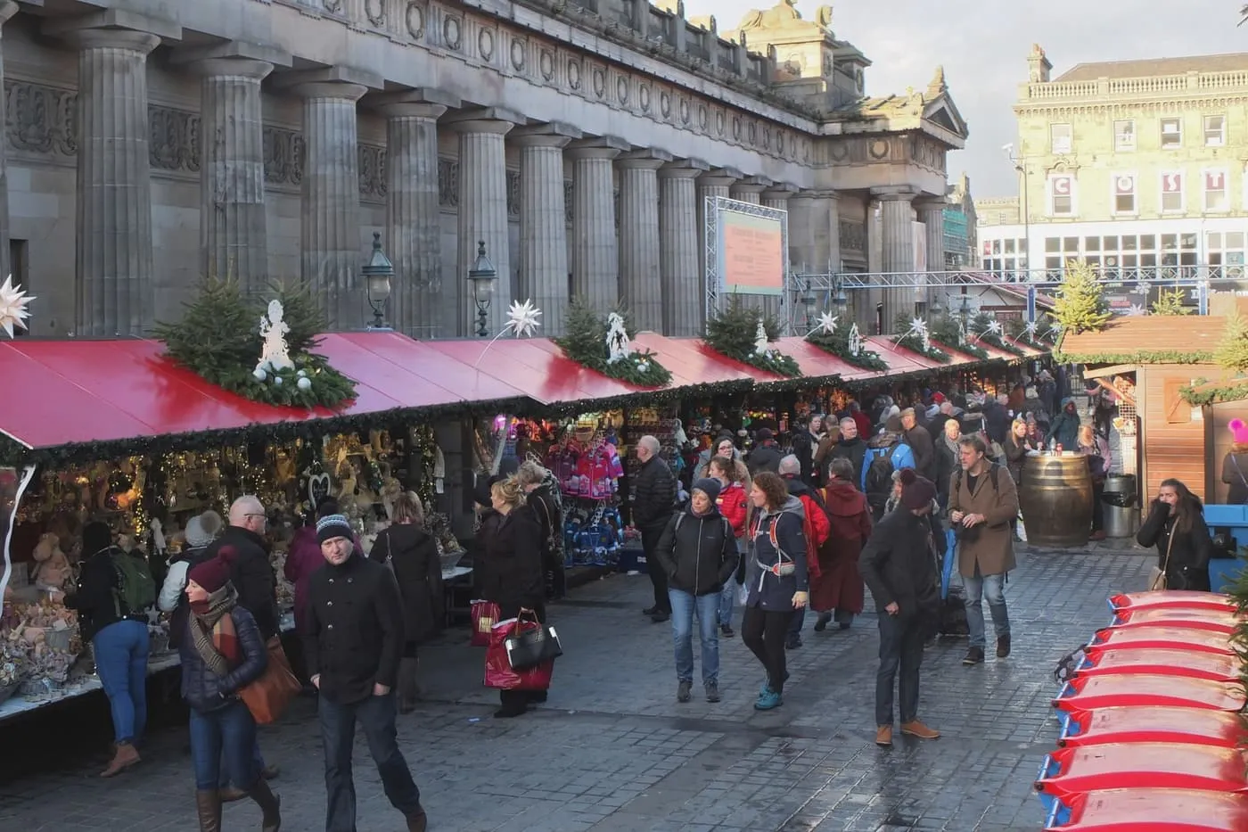 Red-roofed market stalls trimmed with greenery and stars, with shoppers in winter coats walking past and a stone building with a row of tall columns behind