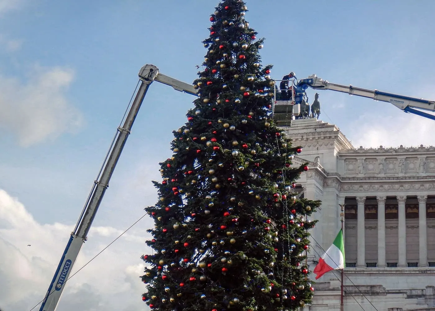 A tall Christmas tree in Piazza Venezia being dressed with baubles by a crane, with the white Vittoriano behind it
