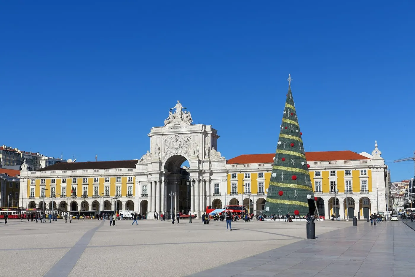 A tall Christmas tree in Praça do Comércio under a clear blue sky, beside the Rua Augusta Arch and the yellow arcades