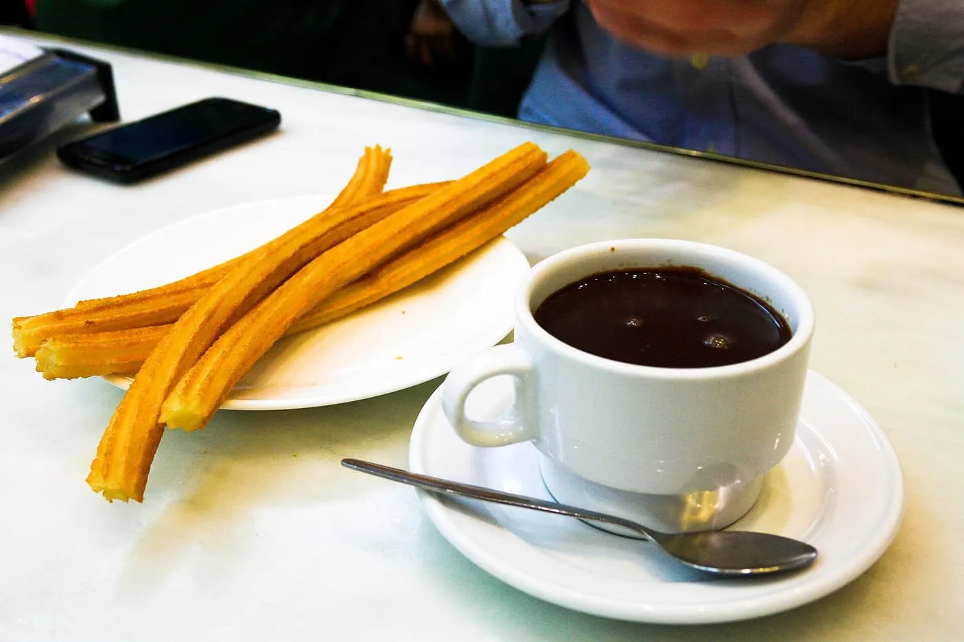 Three churros on a small plate beside a cup of thick hot chocolate on a marble counter