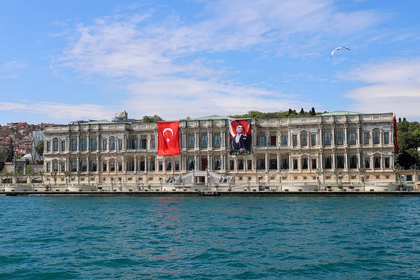 The long white marble facade of Çırağan Palace on the Bosphorus shore, with Turkish flags hanging from it