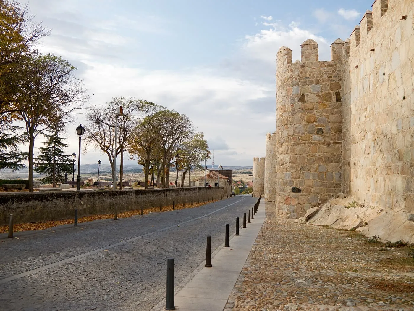 A round tower and the granite wall of Ávila beside a cobbled road lined with bollards and autumn trees