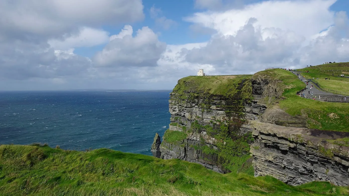 A grass-topped cliff of layered dark rock above the Atlantic, with O'Brien's Tower small on the skyline