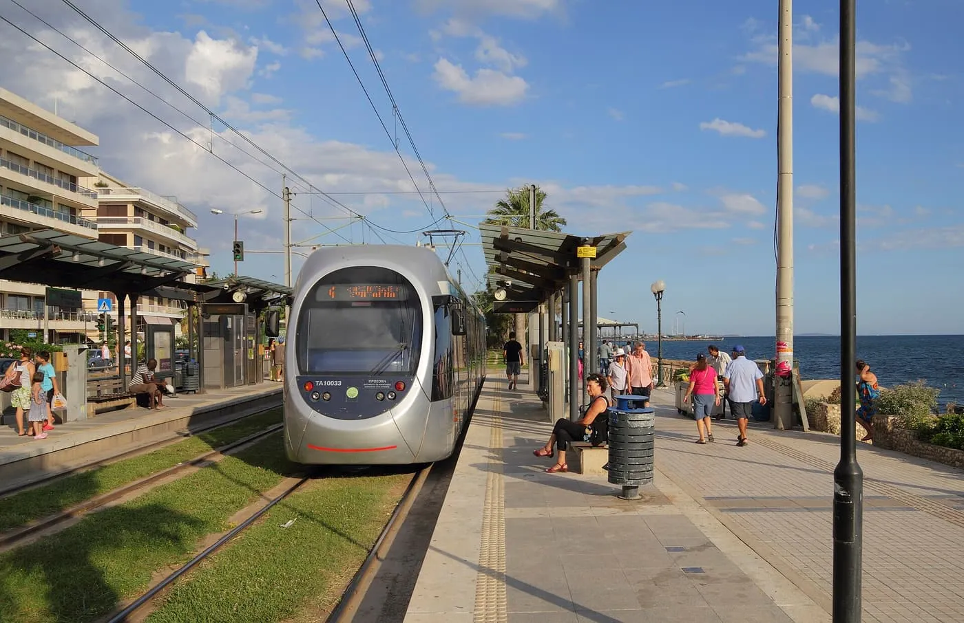 A silver Athens tram standing at a seafront stop on the coast at Paleo Faliro, with the sea beside the platform