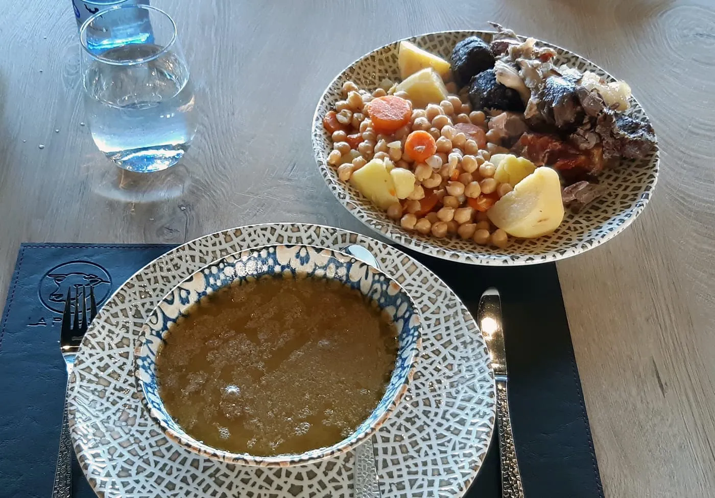 A bowl of cocido broth beside a plate of chickpeas, potato, carrot and meats on a wooden table