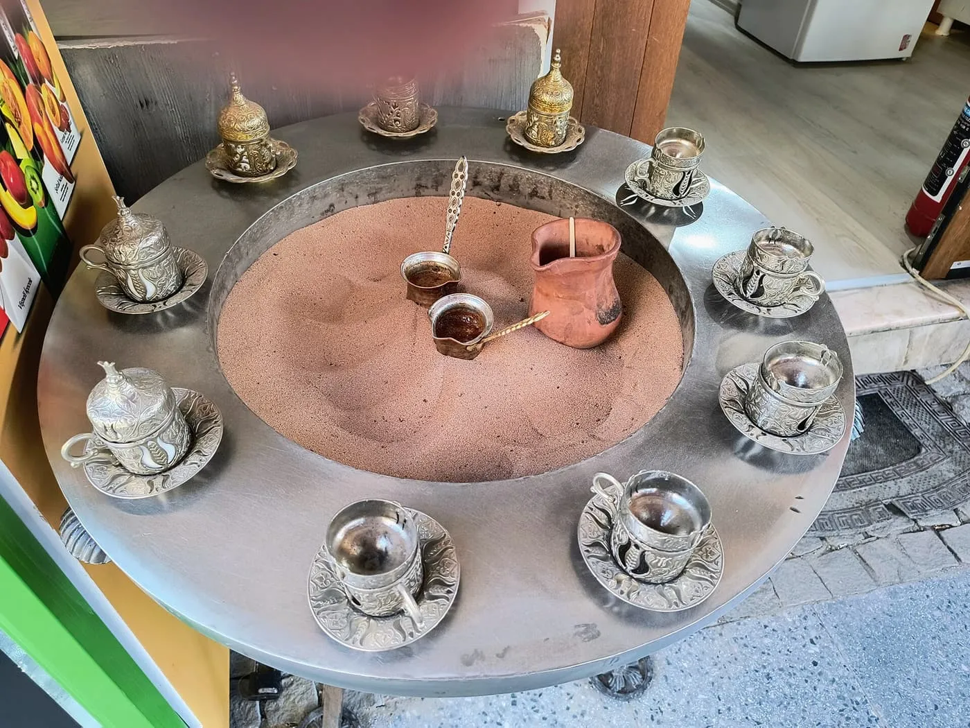 Two small copper cezves standing in a tray of hot sand surrounded by cups on saucers