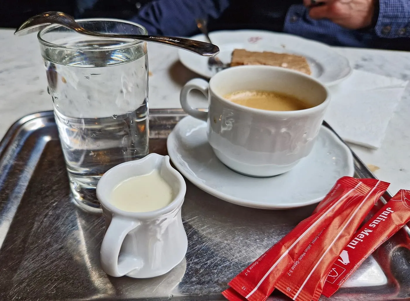 A cup of coffee with a glass of water and a jug of cream on a metal tray on a marble table