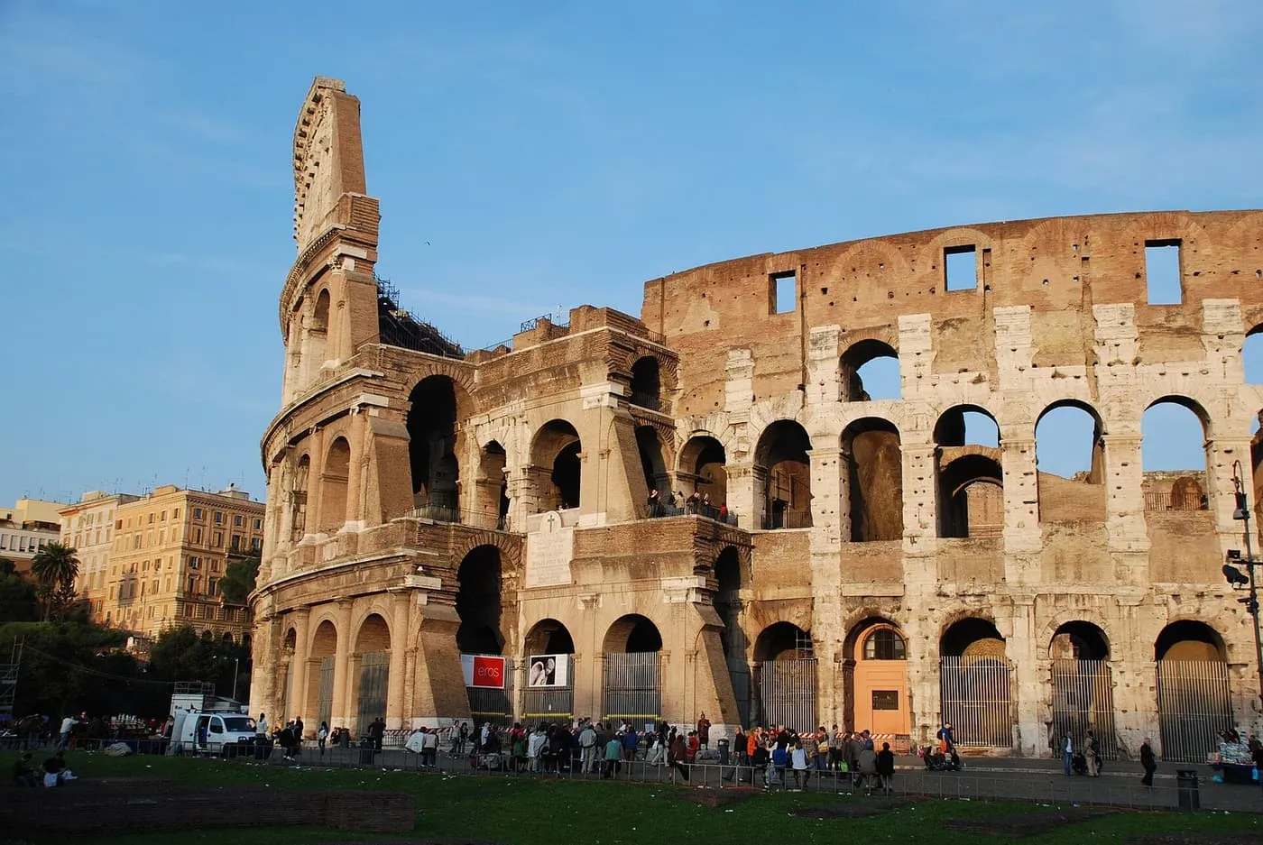 The Colosseum's outer wall and arches under a blue sky in late-afternoon light, with small groups of visitors at its foot