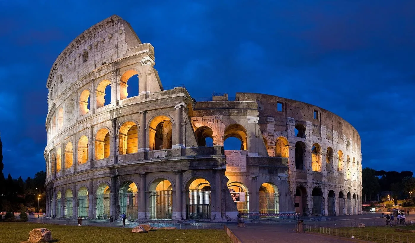 The Colosseum at dusk, its arches lit from within, with a few visitors at its foot