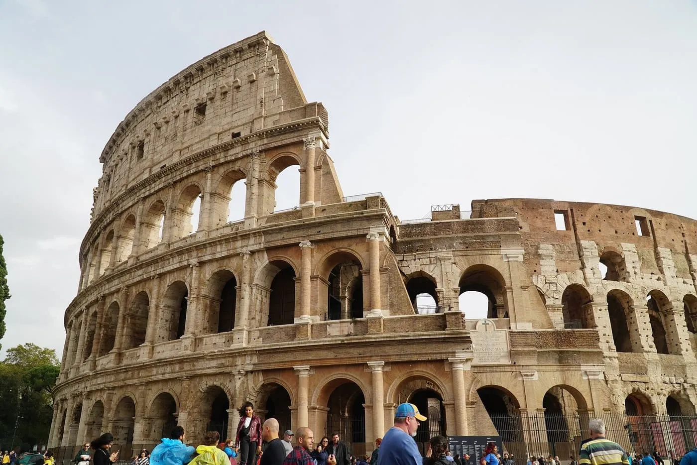 The Colosseum from outside, with visitors gathered along the fence at its foot under a pale sky