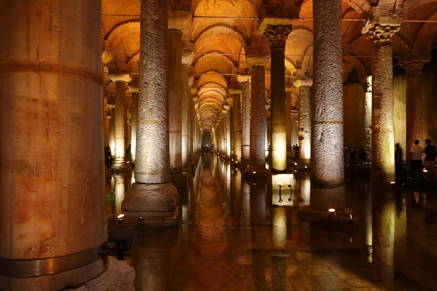 Rows of lit stone columns receding down the Basilica Cistern, with a column in the foreground and water reflecting the lights