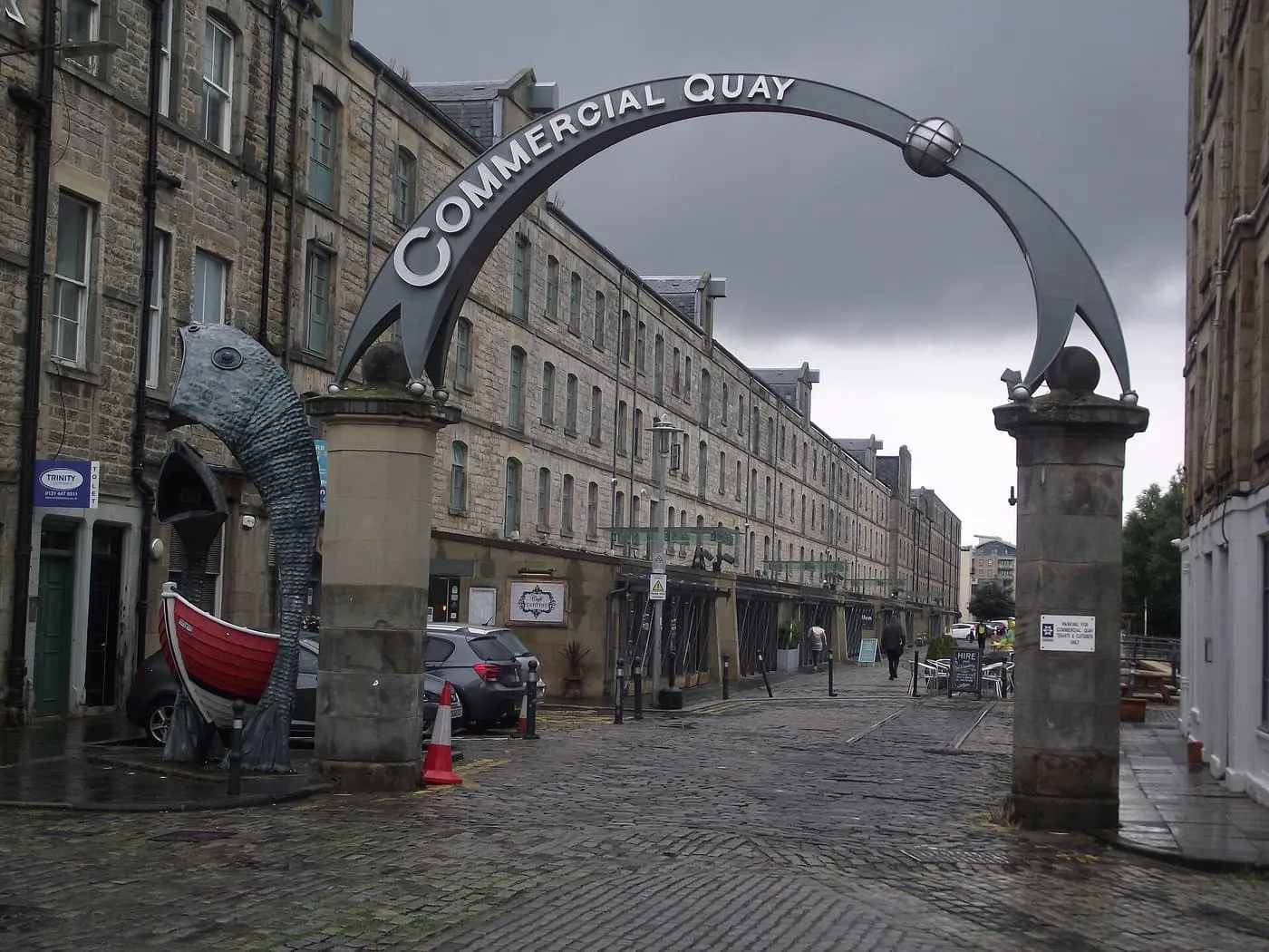 A steel arch reading Commercial Quay over a cobbled Leith street, with a fish sculpture beside one pier and a long row of stone warehouses beyond