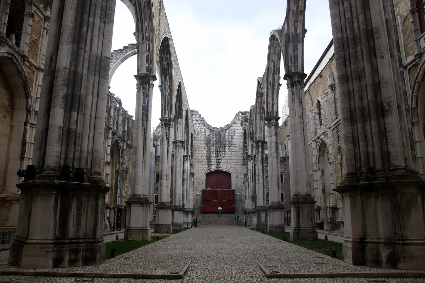 The nave of the Carmo Convent church, roofless, with its tall pointed arches and a cobbled aisle leading to the apse under a pale sky