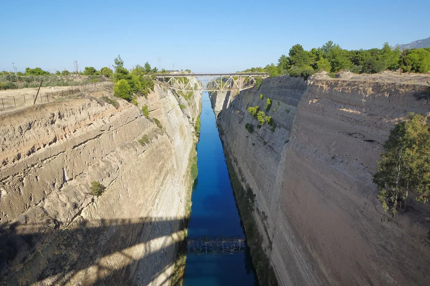 The Corinth Canal seen from above, a strip of blue water between pale limestone walls with a bridge across the far end