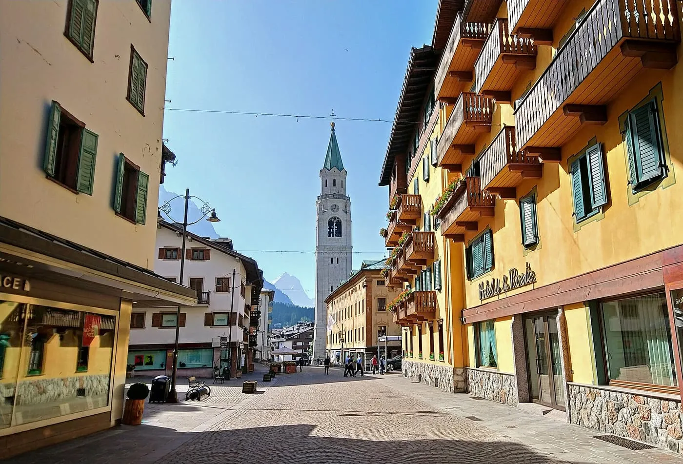 A pedestrian street of ochre buildings with wooden balconies in Cortina d'Ampezzo, with a pale bell tower at the end and mountains behind