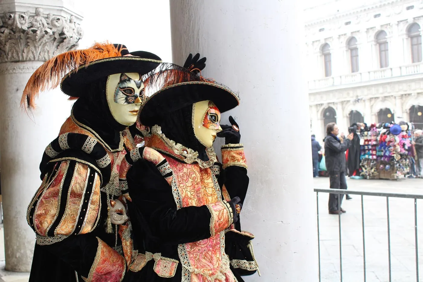 Two people in black velvet and orange brocade costumes with feathered hats and white masks beside a white stone column