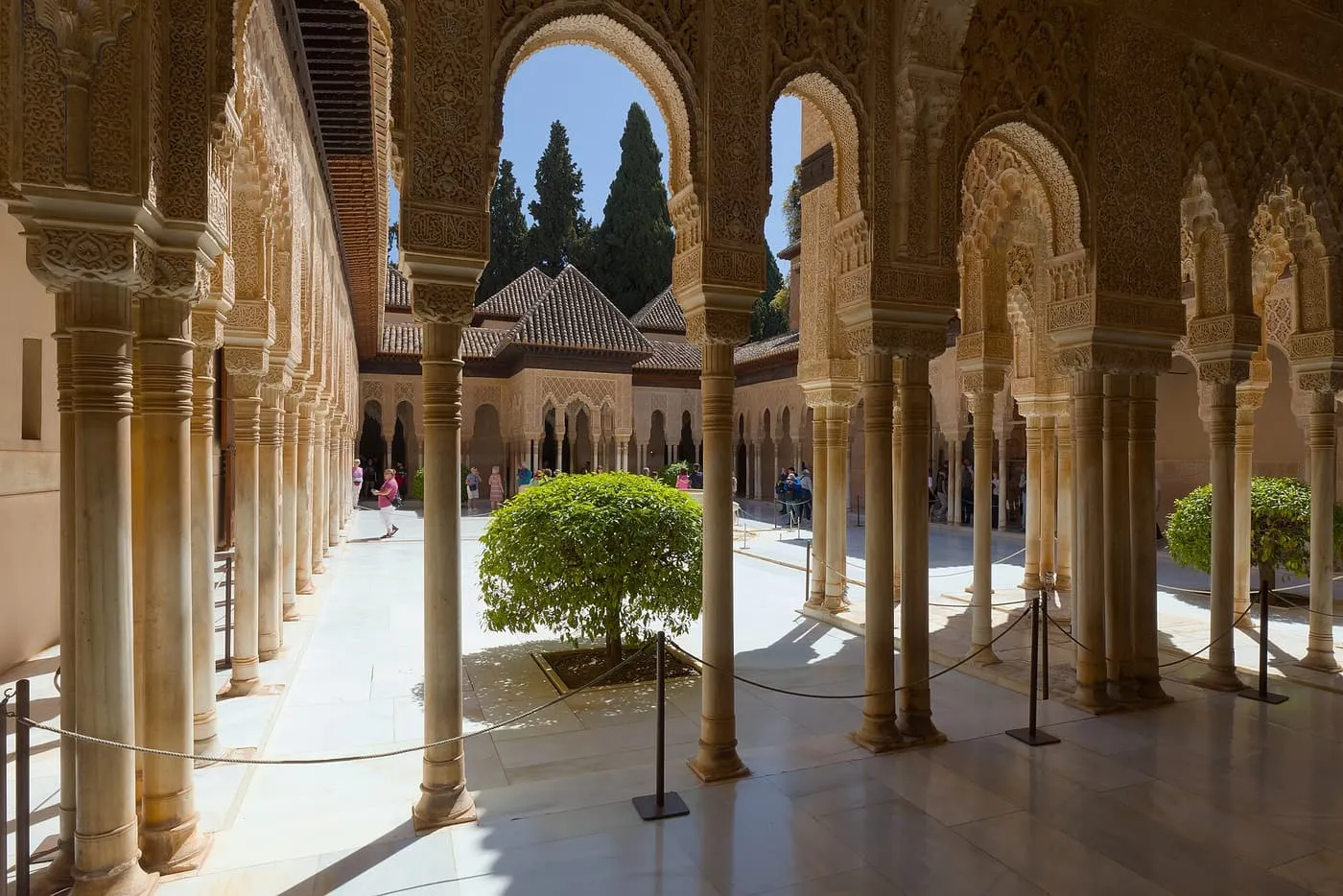 The Court of the Lions in the Alhambra seen through a gallery of slender columns and carved arches, with an orange tree in the courtyard