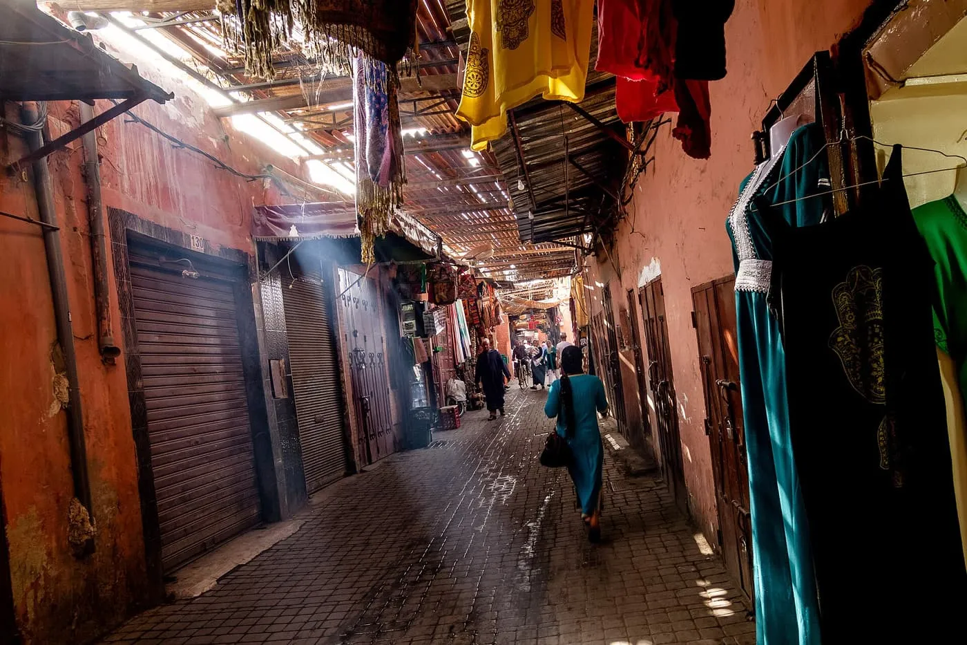 A covered souk lane with shafts of daylight falling through the slatted roof onto the paving, and clothes hanging at the right