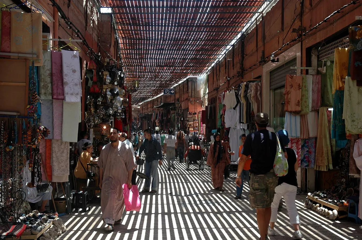 A covered souk lane with a slatted roof casting stripes of light, shops hung with cloth on both sides and shoppers walking along it