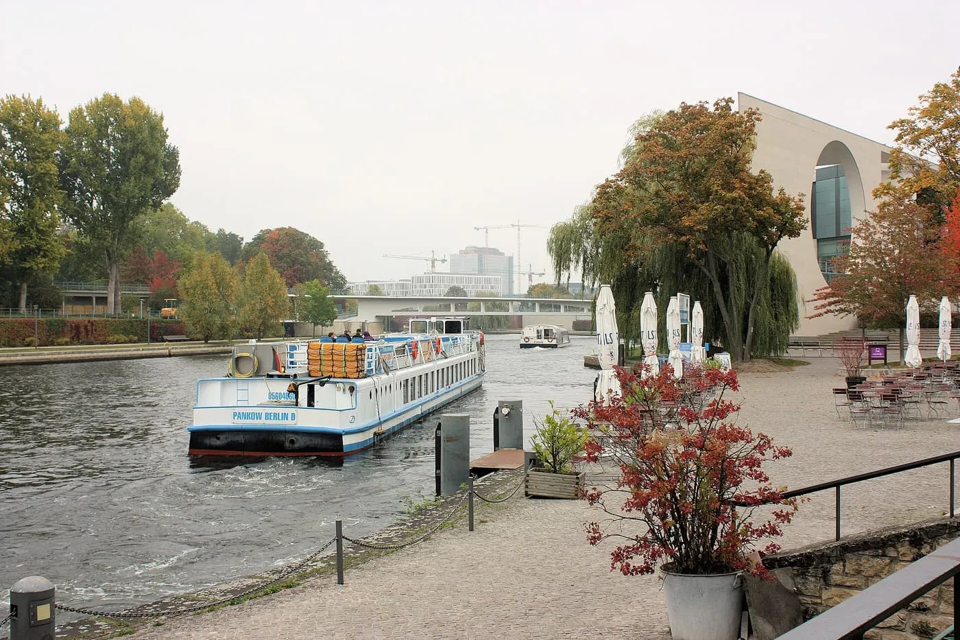 A white excursion boat on the Spree beside the curved stone wall of the Federal Chancellery, with autumn trees and a footbridge beyond