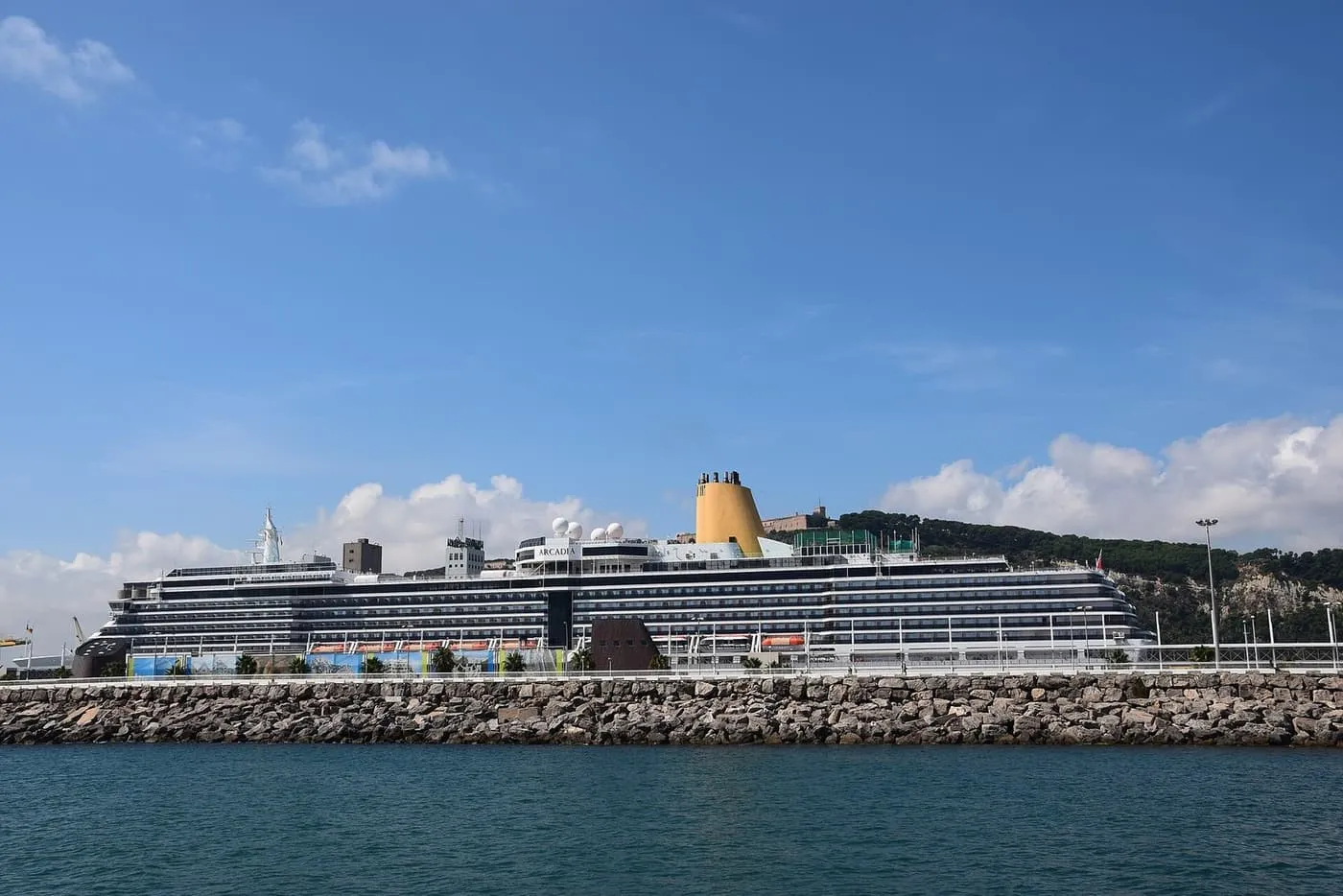 A cruise ship with a yellow funnel moored along a quay in the Port of Barcelona, with the wooded hill of Montjuïc and its castle behind