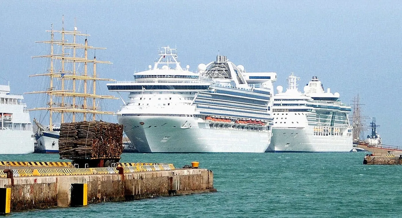 Two large cruise ships moored side by side at the port of Civitavecchia, with a tall ship's masts behind them