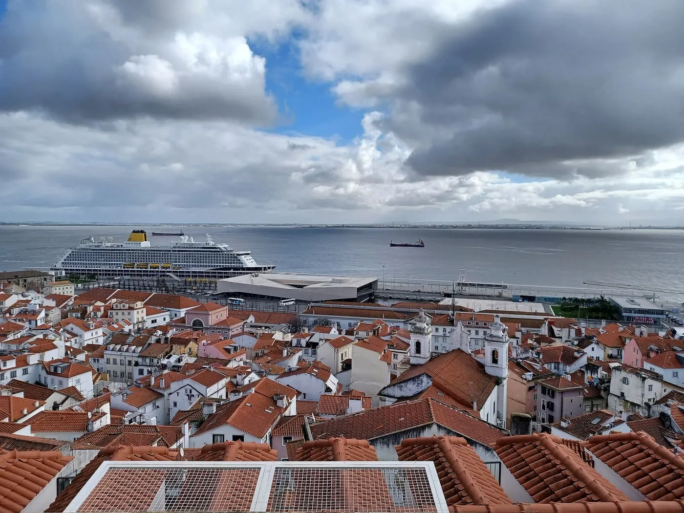 A cruise ship alongside the pale Lisbon cruise terminal on the Tagus, seen over the red roofs of Alfama